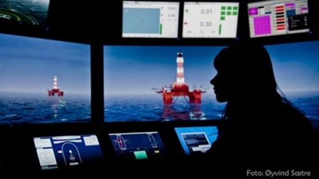 En person sitter ved kontrollpanelet og overvåker oljeplattformen i havet. Skjermene viser ulike data og grafikk.
