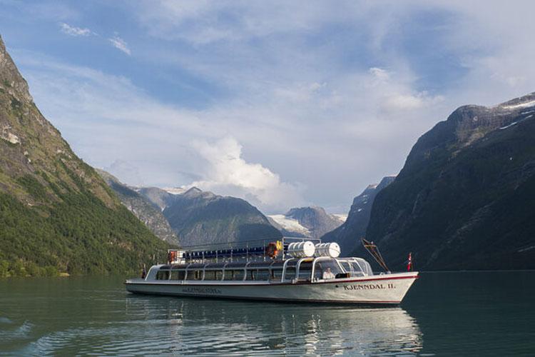 En passasjerbåt seiler på en rolig fjord, omringet av majestetiske fjell og grønn vegetasjon.