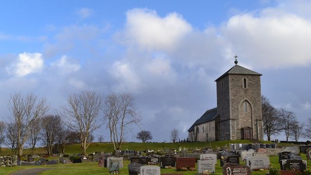 En steinkirke med klokketårn omgitt av gravsteiner under en blå himmel med skyer.