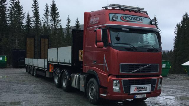 Rød lastebil fra Volvo med flatbed og trelast på tilhenger, parkert i skogsområde med søppeldunker i bakgrunnen.