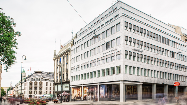 Moderne bygning i Oslo med glassfasade og nærliggende historiske bygninger. Gateutsikt med folk og trafikk.