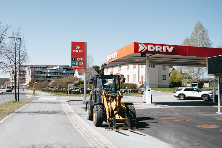 En gravemaskin står parkert ved en DRIV bensinstasjon med prisskilt i bakgrunnen.