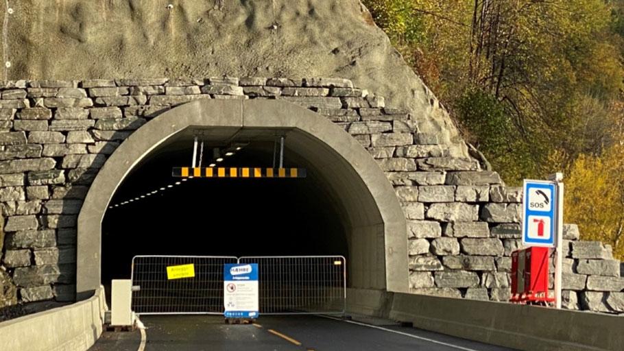 Tunnelåpning med steinvegg og avsperring, skilt som viser veibeskrivelse og advarsel.
