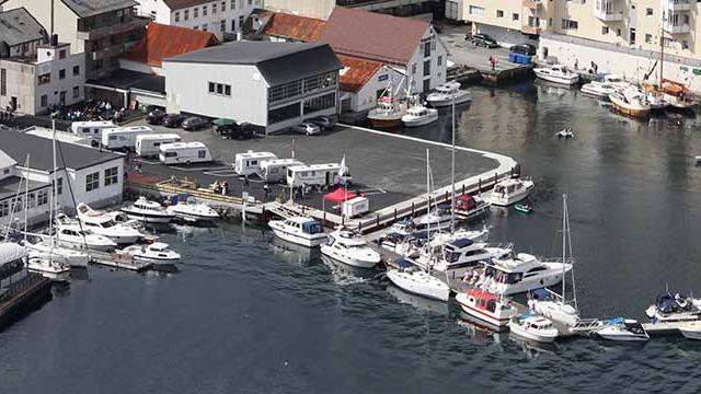 Båthavn med flere båter og campingvogner ved vannkanten, omgitt av bygninger og en rolig havn.