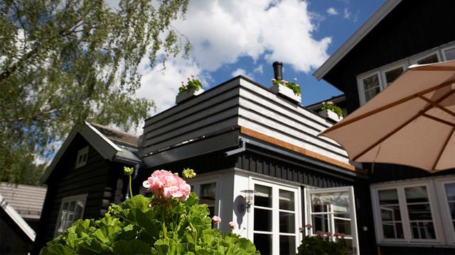 Hus med svart og hvit fasade, blomsterkasser på taket og parasoll i hagen under en blå himmel med skyer.