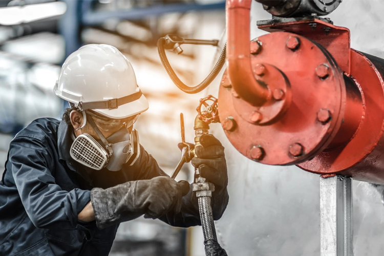 En arbeidstaker i hjelm og verneutstyr jobber med rør og ventiler i en industriell setting.