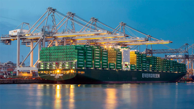 Containerbåt med grønne containere ved havnen, opplyst av lys i skumringen. Kraner i bakgrunnen.