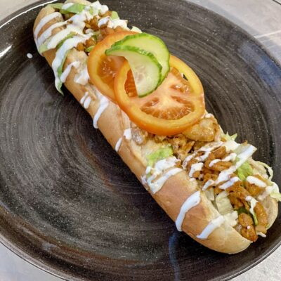 En pølse med kylling, salat, tomat og agurk, servert i et brød med hvit saus på en svart tallerken.