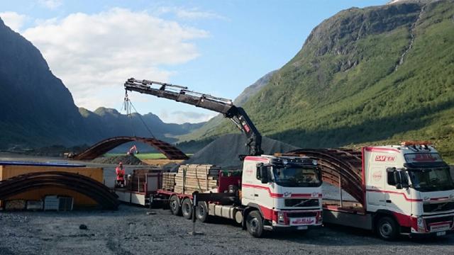 To lastebiler med kran løfter materialer på en byggeplass i fjellandskap.