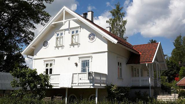 Et hvitt hus med rød takstein, balkong og store vinduer, omgitt av trær og blå himmel med skyer.