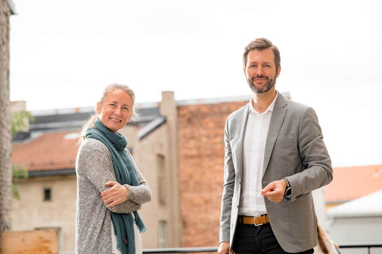 To personer smiler og står sammen utendørs, med bygninger i bakgrunnen. En kvinne med skjerf og en mann i dress.