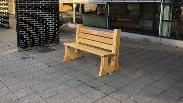 En trebenk plassert på flislagt fortau foran en moderne bygning med glassfasade.