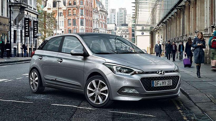 En sølvfarget Hyundai bil parkert på en gate med bygninger og mennesker i bakgrunnen.
