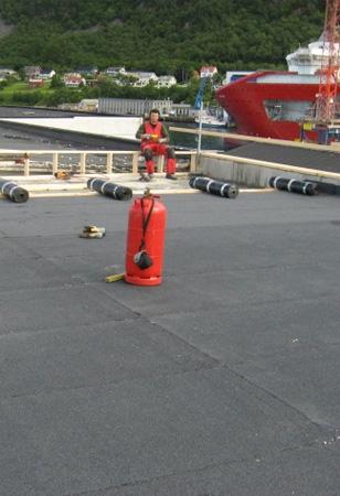 En rød bøye står på en kaiplattform, med en person i arbeid i bakgrunnen og båter i havnen.