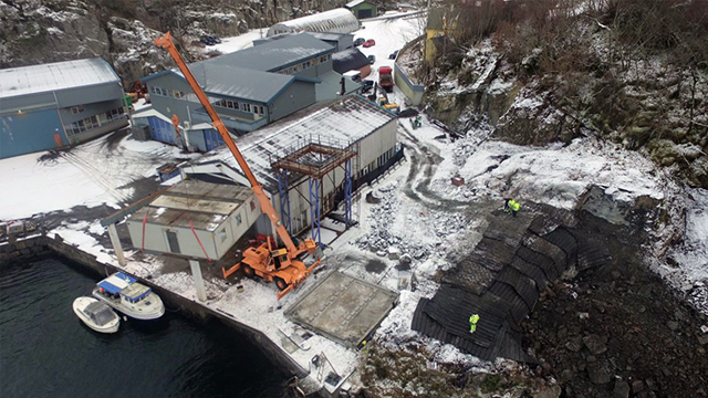 Byggeplass ved havet med kran, båter og snødekte områder rundt bygninger. Arbeidere jobber på stedet.