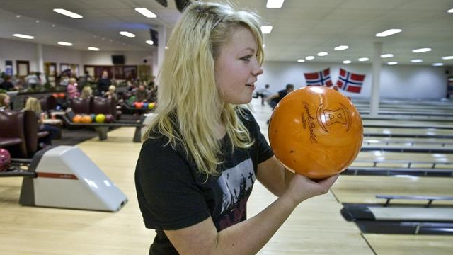 En jente holder en oransje bowlingkule i en bowlinghall med folk i bakgrunnen.