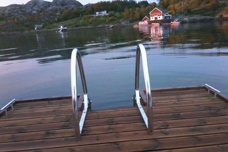 Badebrygge med stige ved vannet, med en rød hytte i bakgrunnen og fjell i horisonten.