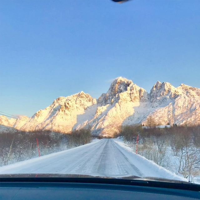 Vintervei med snødekte fjell i bakgrunnen under en klar blå himmel.