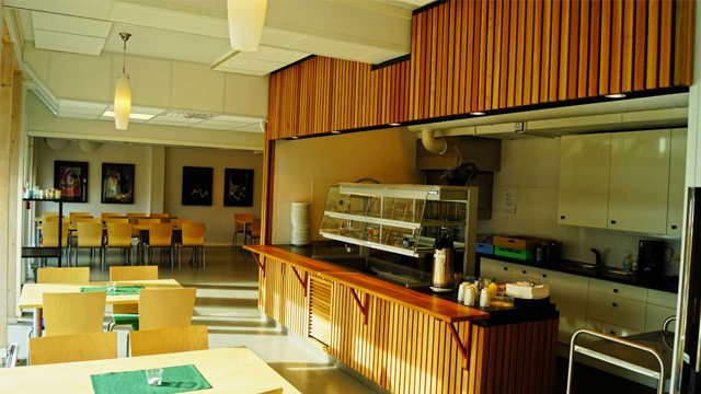 Kafé med trepanel, bord og stoler, samt en serveringsdisk med kaffemaskin og kjøkkenutstyr.