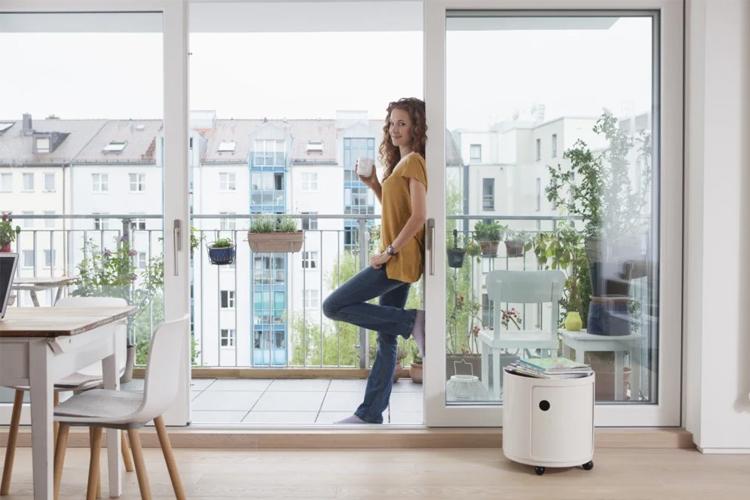 Kvinne står på balkongen med en kopp i hånden, omringet av planter og moderne bygninger i bakgrunnen.