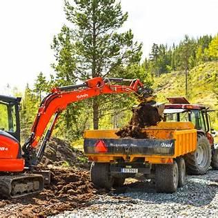 Gravemaskin laster jord i en lastebil på en byggeplass i skogen, omringet av trær og grønt landskap.