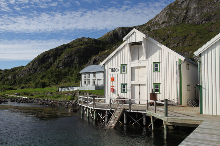 En hvit bygning ved vannkanten med en brygge, omgitt av grønne fjell og blå himmel.