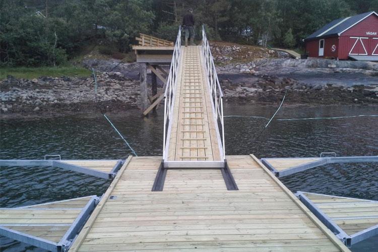 Bilde av en trebro som fører til en liten kai ved vannet, med en rød hytte i bakgrunnen.