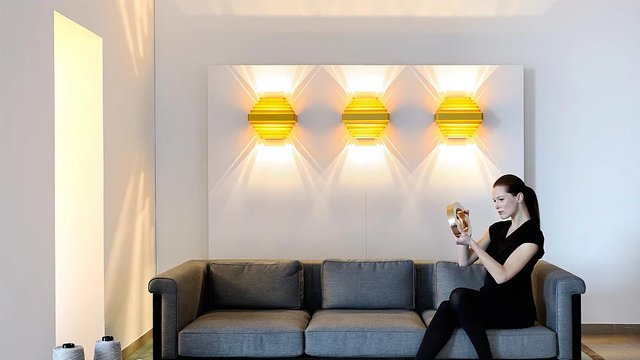 En kvinne sitter på en grå sofa og holder en gjenstand, med tre gule vegglamper i bakgrunnen.