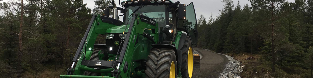 Grønn traktor med frontlaster på en grusvei omgitt av trær i skogen.
