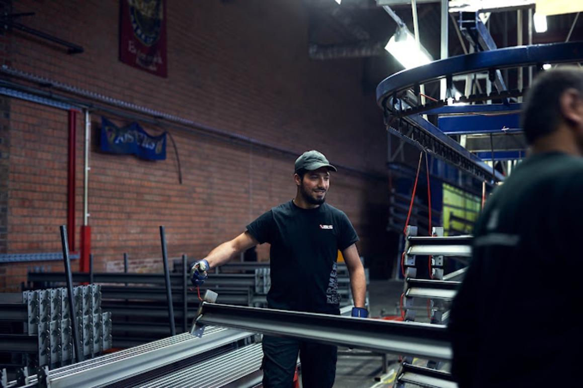 En mann i svart t-skjorte jobber med metallstenger i en fabrikk, omgitt av maskiner og utstyr.