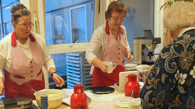 To kvinner i forkleer serverer mat og drikke til en eldre kvinne i et lyst kjøkkenmiljø.