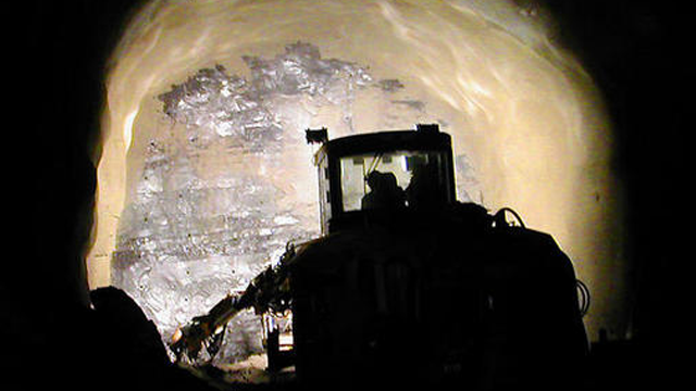 Gravemaskin som arbeider i en tunnel med lys som belyser veggen av fjell.