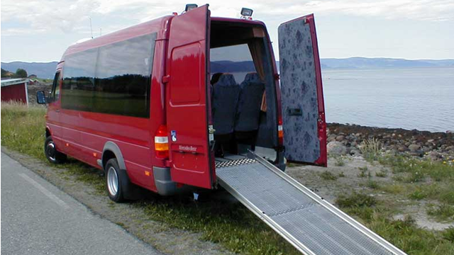 Rød minibuss med rampe, parkert ved sjøen, med åpne bakdører og seteinnredning synlig.