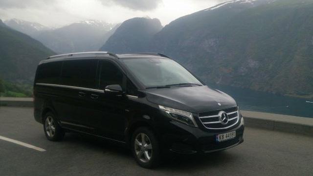 En svart Mercedes varebil parkert ved en fjord med fjell i bakgrunnen.
