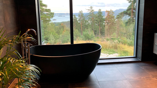 Moderne bad med svart badekar og stor vindu som viser utsikt over skog og innsjø.