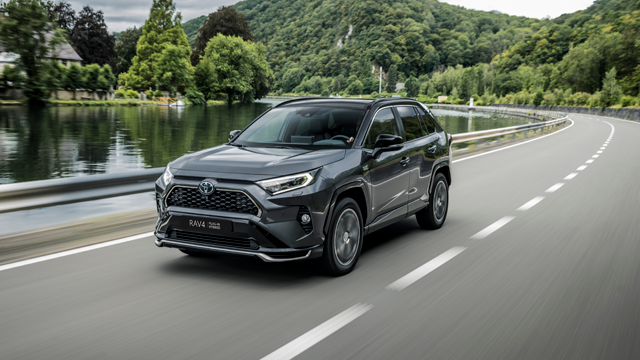 En grå Toyota RAV4 SUV kjører langs en vei ved en elv, omkranset av grønne trær og fjell.