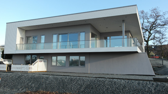 Moderne hus med stor terrasse og glassrekkverk, beliggende på en steinete tomt.
