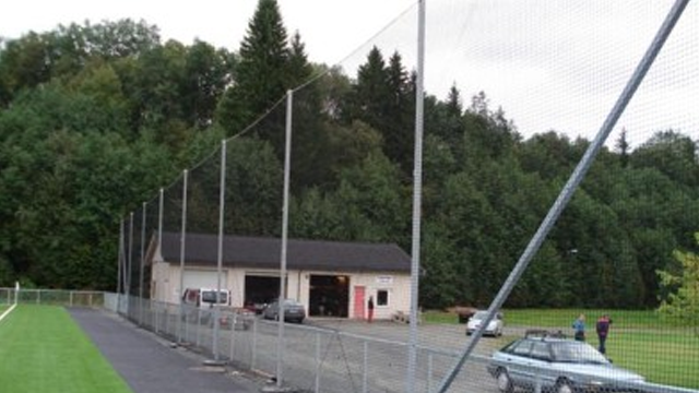 En fotballbane med et klubbhus og høye nett rundt, omgitt av trær og parkerte biler.