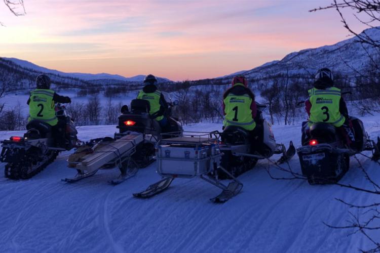 Fire snøscootere med førere i gule vester, som ser på solnedgangen over fjellene.