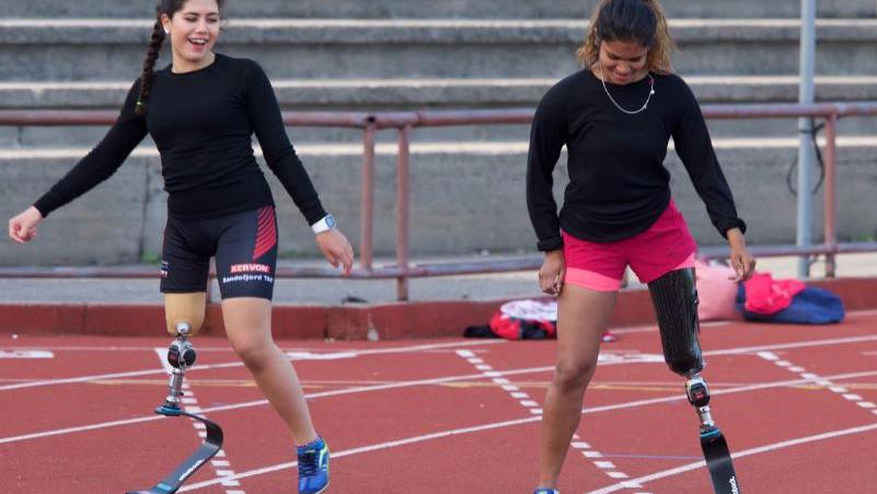 To kvinner med proteser trener på en løpebane, iført sportsklær og smilende.
