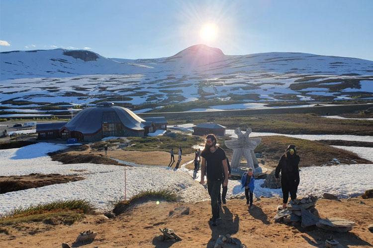 Folk går nedover en sti i snødekt landskap med en bygning og fjell i bakgrunnen under soloppgang.