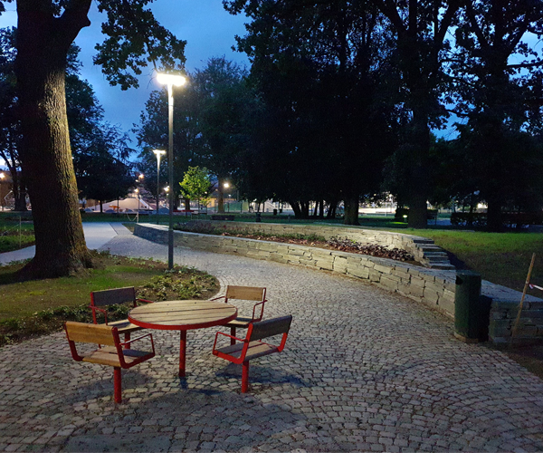 Et bord med stoler i en park om kvelden, omgitt av trær og belysning langs en brosteinssti.