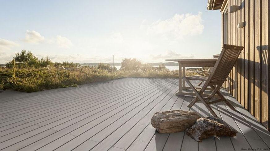Utsikt fra en terrasse med sollys, stoler, bord og steiner, omkranset av grønt gress og hav i bakgrunnen.
