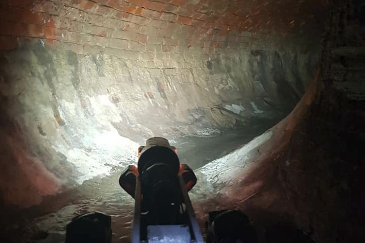 Kamera som viser en buet murvegg i en underjordisk tunnel med svakt lys.