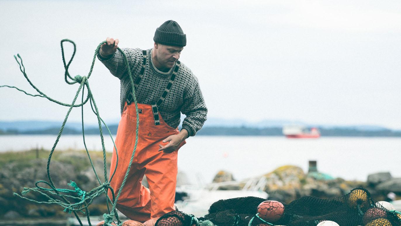 Fisker i oransje fiskeutstyr som håndterer garn ved sjøen, med en båt i bakgrunnen.