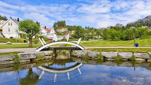En vakker bro over en elv med speilbilde, omgitt av grønne trær og hus i en naturskjønn landsby.
