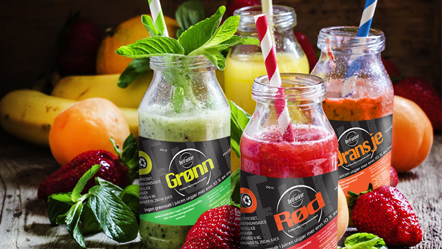 Fire smoothie-flasker med fargerike drikker, omringet av frukt og mynteblader på en treoverflate.