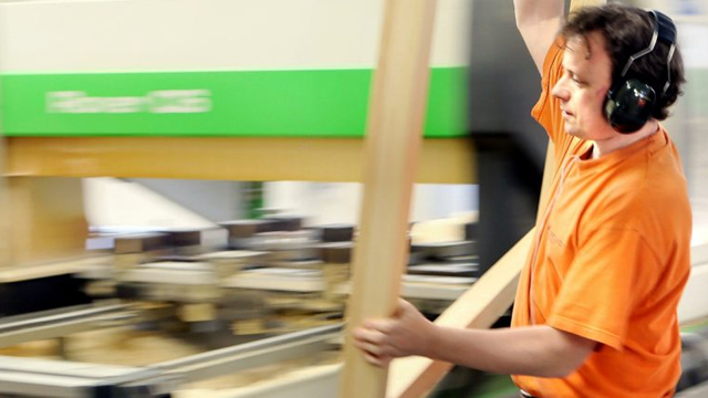 En mann i oransje t-skjorte og hodetelefoner jobber med trebearbeiding i en fabrikk.