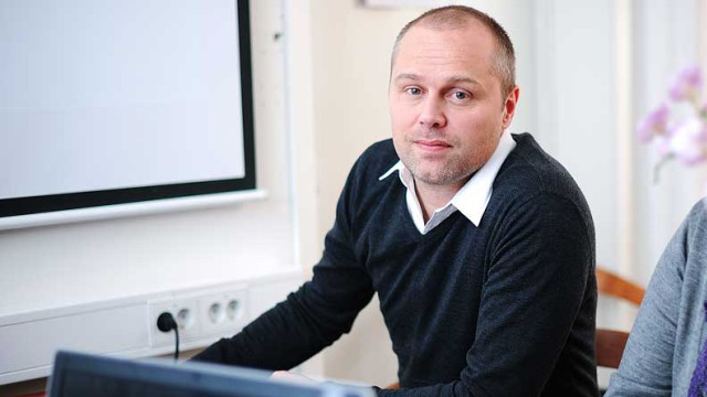 Mann i svart genser sitter ved skrivebord med datamaskin, ser mot kamera i kontormiljø.