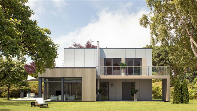 Moderne hus med stor hage, glassfasade og balkong, omgitt av trær og grøntområder.